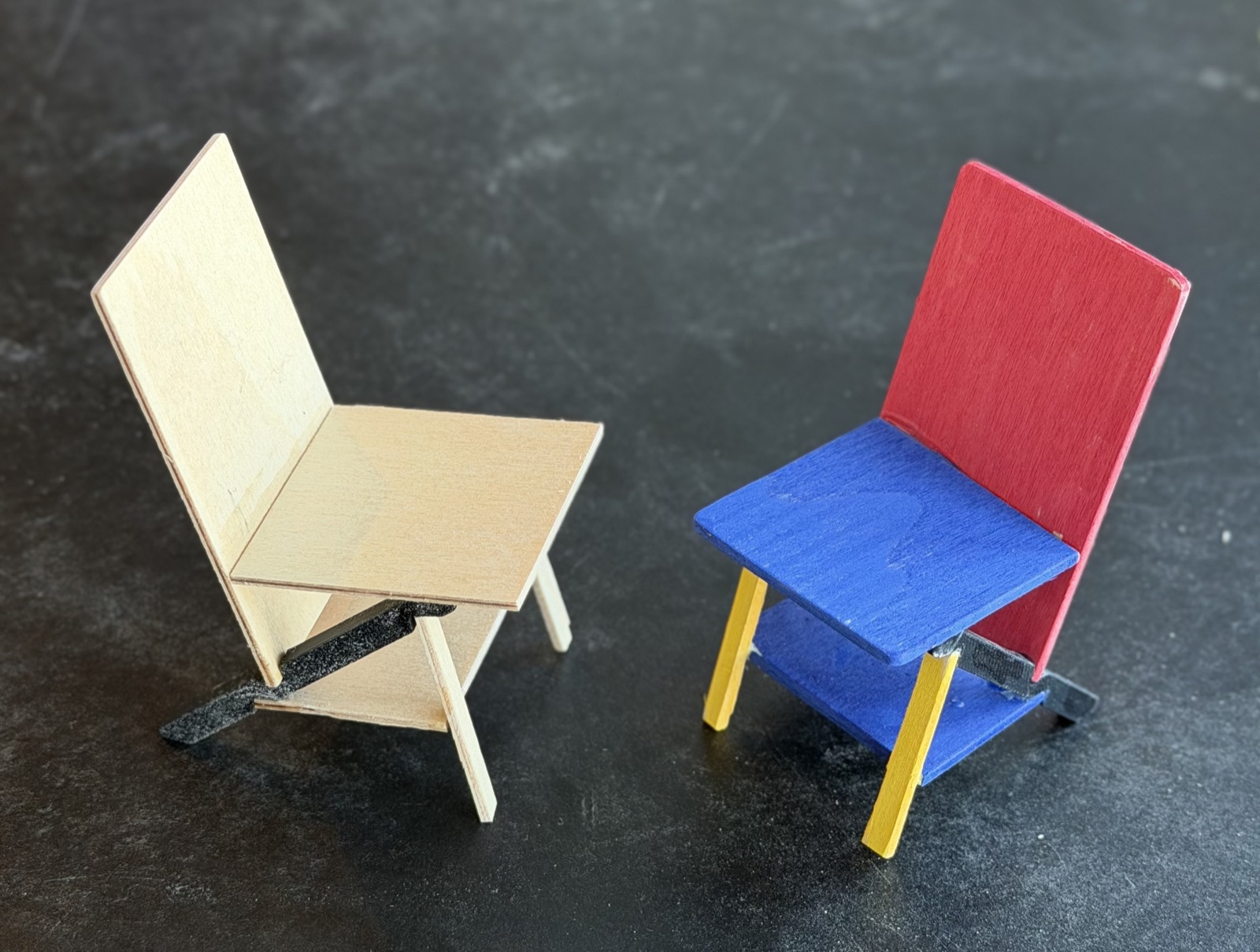Two small plywood chair models on a dark surface, one natural, one with a red back, blue seat and yellow legs
